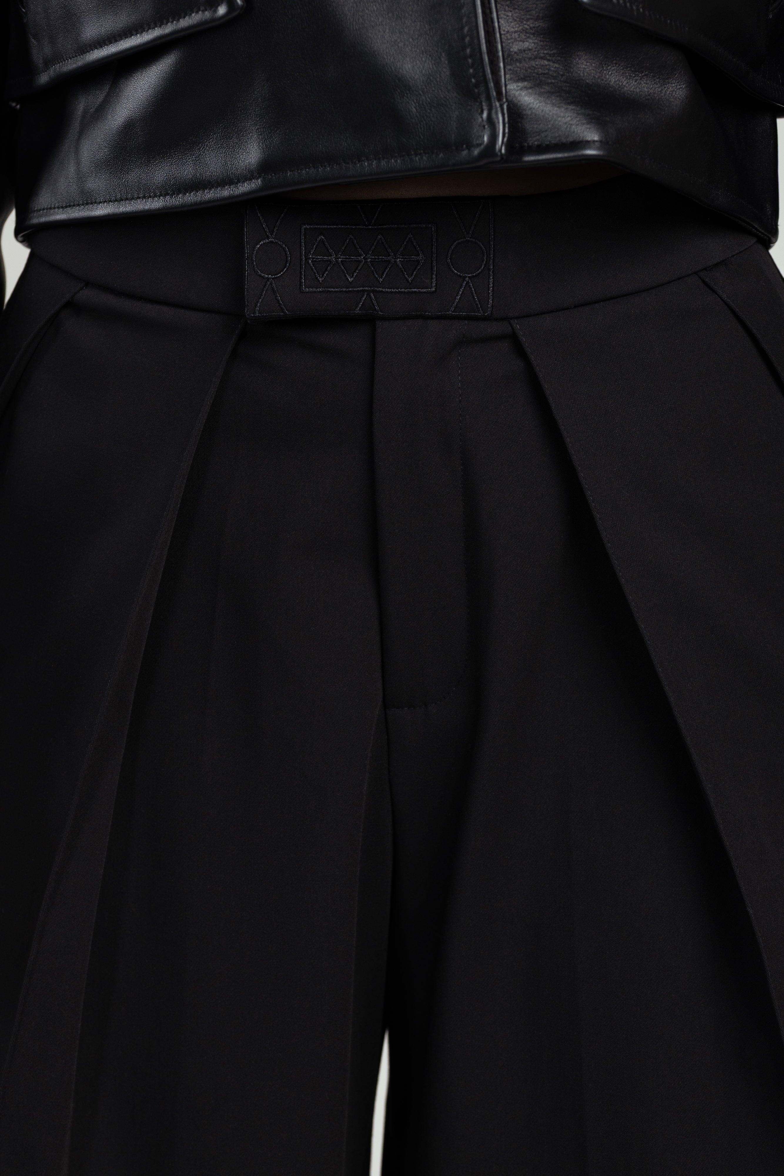 Close-up of a black pleated pants with an embroidered brand logo on a white background